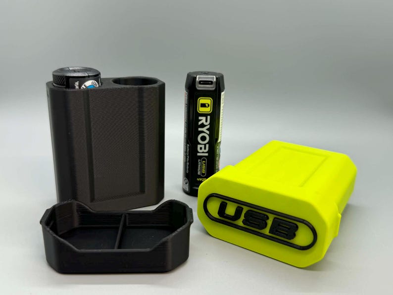 Op de afbeelding: Een zwarte 3D-geprinte batterijhouder met twee sleuven, een bijpassende zwarte basisplaat, een limoengroene batterijafdekking met het woord USB in reli&euml;f in het zwart, en een zwarte Ryobi USB-lithiumbatterij die tussen de houder en de afdekking staat.