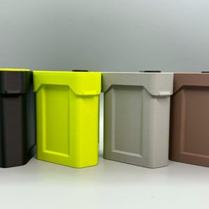 Op de afbeelding: Vier rechthoekige opbergbakken op een rij, met een matte afwerking en een verzonken paneelontwerp. De bakken zijn zwart, neongeel, lichtgrijs en bruin van kleur, elk voorzien van een klein zwart vierkant deksel aan de bovenzijde.