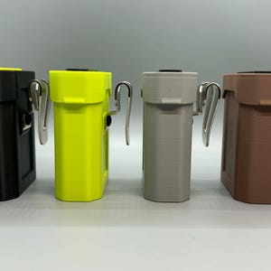 Op de afbeelding: Vier rechthoekige riemcliphouders op een rij, in de kleuren zwart, neongeel, lichtgrijs en bruin. Elke houder is voorzien van een verticale metalen clip aan de zijkant en een plat bovenoppervlak, uitgevoerd met een matte textuur.
