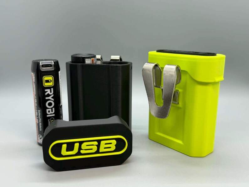 Op de afbeelding: Een zwarte Ryobi lithiumbatterij, een zwarte cilindrische stroomadapter, een zwarte rechthoekige afdekking met de gele tekst USB, en een fel limoengroene stroomadapterbehuizing met een zilveren metalen riemclip aan de zijkant.