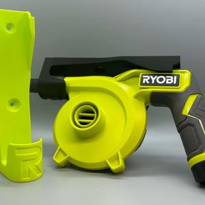 May include: A lime green Ryobi handheld power blower with a black handle and top housing, positioned next to a matching lime green wall-mounted storage bracket featuring a raised letter R on the front base and two circular screw holes.