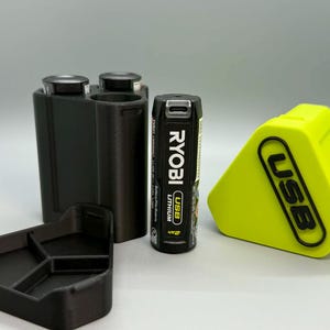 Op de afbeelding: Een zwarte 3D-geprinte batterijhouder met twee ronde sleuven, een aparte zwarte driehoekige basis, een zwarte Ryobi USB-lithiumbatterij en een fel neongele driehoekige hoes met de in reli&euml;f aangebrachte zwarte tekst USB op de voorzijde.