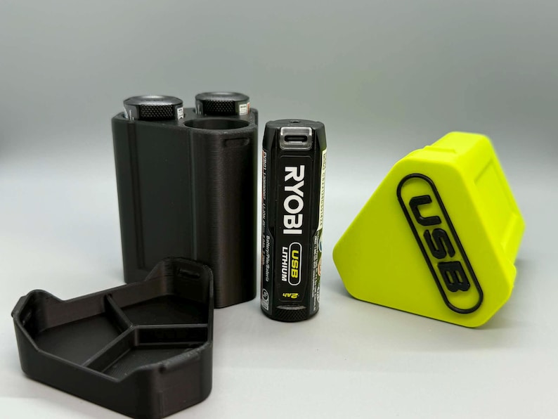 Op de afbeelding: Een zwarte 3D-geprinte batterijhouder met twee ronde sleuven, een aparte zwarte driehoekige basis, een zwarte Ryobi USB-lithiumbatterij en een fel neongele driehoekige hoes met de in reli&euml;f aangebrachte zwarte tekst USB op de voorzijde.