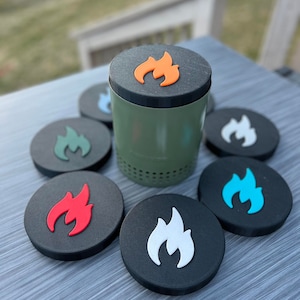 May include: A green metal container with a black lid featuring an orange flame design. Surrounding the container are several black coasters, each with a different colored flame design, including red, white, blue, and green. The items are arranged on a gray surface.