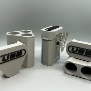 Op de afbeelding: Een collectie lichtgrijze 3D-geprinte opbergorganizers voor USB-sticks. De set bevat een driehoekige houder, een rechthoekige behuizing met een metalen clip en een houder met twee sleuven. Elk stuk heeft een zwart label met de tekst USB.