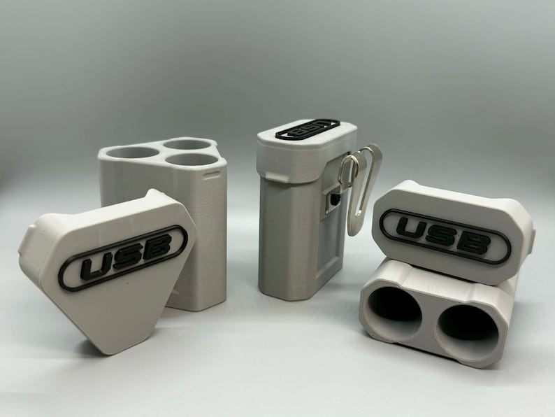 Op de afbeelding: Een collectie lichtgrijze 3D-geprinte opbergorganizers voor USB-sticks. De set bevat een driehoekige houder, een rechthoekige behuizing met een metalen clip en een houder met twee sleuven. Elk stuk heeft een zwart label met de tekst USB.
