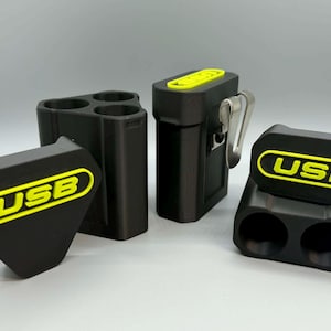 Op de afbeelding: Een set zwarte opbergdoosjes voor USB-sticks met felgele accenten en de tekst USB. De doosjes bevatten een driehoekige houder met drie sleuven, een rechthoekige houder met &eacute;&eacute;n sleuf en een metalen riemclip, en een houder met twee sleuven en deksel.
