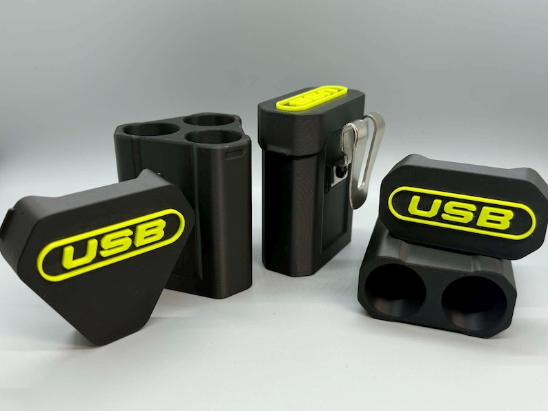 Op de afbeelding: Een set zwarte opbergdoosjes voor USB-sticks met felgele accenten en de tekst USB. De doosjes bevatten een driehoekige houder met drie sleuven, een rechthoekige houder met &eacute;&eacute;n sleuf en een metalen riemclip, en een houder met twee sleuven en deksel.