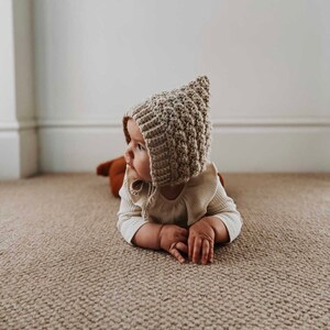 May include: A baby wearing a beige, textured, crocheted bonnet with a pointed top and tie strings. The baby is lying on a beige carpet, wearing a cream-coloured long-sleeved shirt.