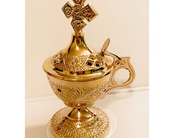 May include: A gold metal censer with a cross on top and intricate floral designs. The censer has a handle and a base with a decorative pattern.