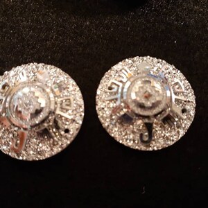 May include: Two silver-toned round metal embellishments with a raised, intricate design. The embellishments have a sparkly, textured surface.