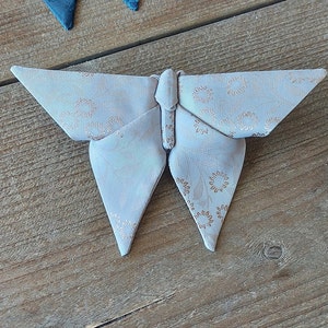 Puede incluir: Una mariposa de origami blanca con un delicado patrón de círculos marrones. La mariposa está doblada de tela y tiene una apariencia tridimensional.