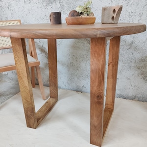 Puede incluir: Una mesa redonda de madera con un acabado de madera natural y un dise&ntilde;o sencillo. La mesa tiene cuatro patas que est&aacute;n hechas del mismo tipo de madera que la superficie de la mesa. La mesa se muestra en una habitaci&oacute;n con una pared blanca y una silla.