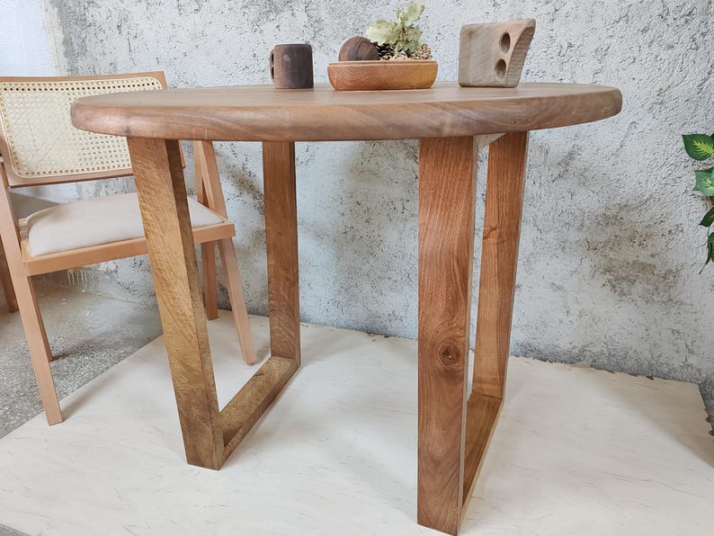 Puede incluir: Una mesa redonda de madera con un acabado de madera natural y un dise&ntilde;o sencillo. La mesa tiene cuatro patas que est&aacute;n hechas del mismo tipo de madera que la superficie de la mesa. La mesa se muestra en una habitaci&oacute;n con una pared blanca y una silla.