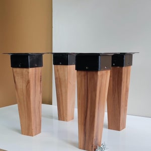 May include: Four wooden table legs with a natural wood grain finish. Each leg has a black metal mounting plate and a tapered design. A set of screws is visible near the legs.