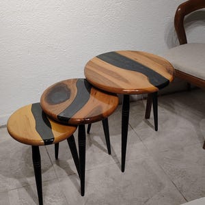 May include: A set of three nesting tables with round wooden tops. The tops have a natural wood grain design with a dark grey resin inlay. Each table has three black, tapered legs. The tables are of varying sizes.