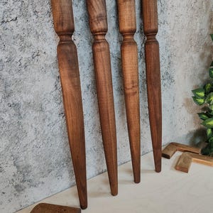 May include: Four wooden furniture legs with a turned design, showcasing a rich brown color and a tapered shape. The legs are standing upright against a textured gray wall, with additional wooden components visible at the bottom. These are likely replacement legs or components for furniture.
