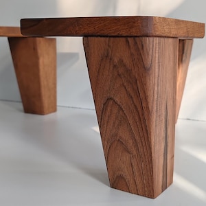May include: A small wooden stand featuring a flat rectangular top supported by four thick, tapered legs. The piece is crafted from natural brown wood with a visible grain pattern, showcasing a smooth finish and sturdy construction throughout.