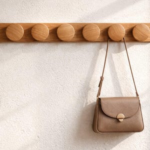 May include: A wooden wall-mounted coat rack with six round pegs. A brown purse with a shoulder strap hangs from one of the pegs. The rack and purse are against a white textured wall.