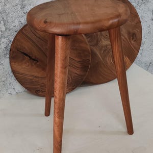 Könnte beinhalten: Ein Holzhocker mit einer runden, braunen Oberseite und drei Beinen. Der Hocker ist aus Naturholz mit rustikalem Finish.