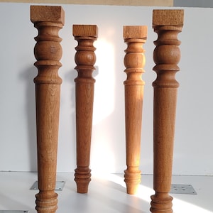 May include: Four light brown wooden furniture legs with a turned, cylindrical design. Each leg features a square top block, a rounded bulbous section near the top, a tapered body, and a small circular foot at the base, standing upright in a row.