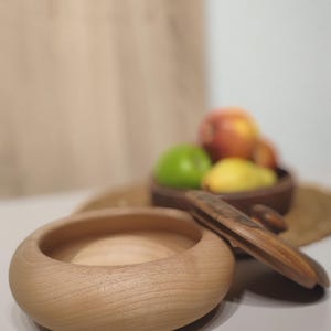 May include: A light-colored wooden bowl with a rounded shape, next to a wooden nutcracker. In the background, a bowl holds green, yellow, and red fruit. The items are on a light-colored surface.