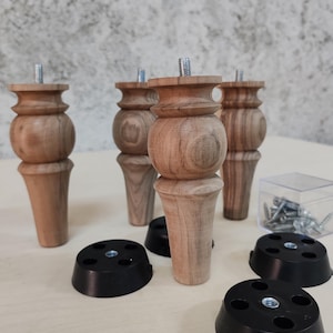 May include: Four turned wooden furniture legs with a natural light brown wood grain finish, each featuring a threaded metal bolt at the top. The set includes four circular black plastic mounting plates and a small clear box containing metal screws.