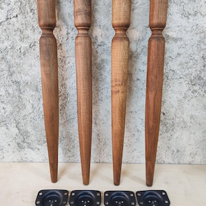 May include: Four wooden furniture legs with a tapered design, each with a cylindrical top and a decorative midsection. The legs are a warm brown color. Included are four black mounting plates and a set of screws.