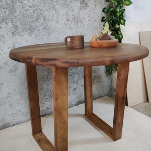 Puede incluir: Una mesa redonda de madera con un acabado de madera natural y un marco de metal rectangular simple. La mesa es peque&ntilde;a y ser&iacute;a adecuada para una mesa auxiliar o una mesita de noche.