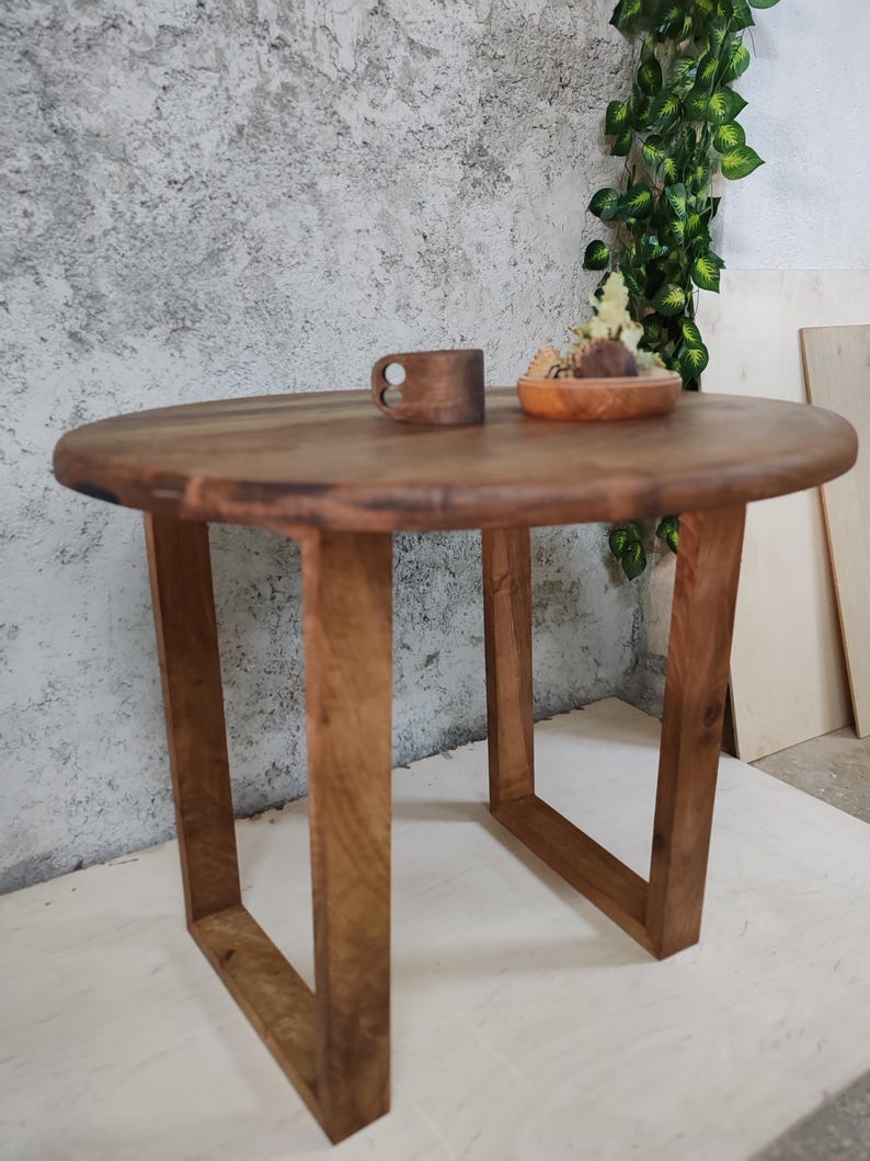 Puede incluir: Una mesa redonda de madera con un acabado de madera natural y un marco de metal rectangular simple. La mesa es peque&ntilde;a y ser&iacute;a adecuada para una mesa auxiliar o una mesita de noche.