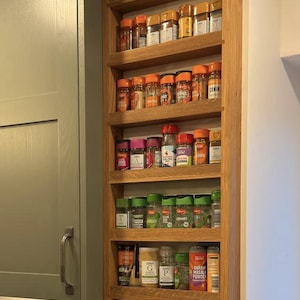 Peut inclure: Un porte-épices en bois avec cinq étagères, monté sur un mur. Les étagères sont remplies de diverses épices dans des bocaux en verre avec des couvercles orange. Le porte-épices est en bois brun clair et a une finition naturelle.
