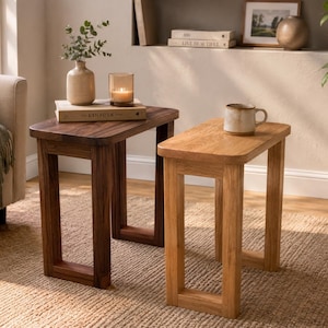 May include: Two rectangular side tables with four-legged frames. One table is crafted from dark brown wood and holds a beige vase, a candle, and a book titled Kinfolk. The second table is made of light oak wood and supports a ceramic mug.