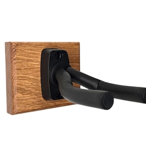 Peut inclure: Un support mural noir pour guitare avec deux bras fixés à une base en bois. Le support est conçu pour maintenir une guitare à l'horizontale.