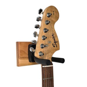 Puede incluir: Un soporte de pared de madera para guitarra con un gancho de metal negro que sostiene una guitarra eléctrica de madera marrón y natural con clavijas de afinación cromadas y el texto "Squier by Fender" en la pala.