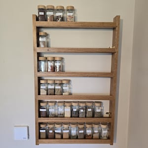 Peut inclure: Un présentoir à épices en bois avec plusieurs étagères, rempli de bocaux en verre contenant diverses épices. Le présentoir est fixé à un mur de couleur claire. Chaque pot a un couvercle en bois et une étiquette.