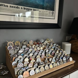 Pode incluir: Um expositor de madeira com várias filas de moedas redondas e coloridas. O expositor é feito de madeira marrom claro e tem um design inclinado. As moedas são dispostas em filas, com cada fila ligeiramente mais alta que a de baixo. As moedas são de diferentes cores e desenhos, e algumas têm um acabamento metálico.