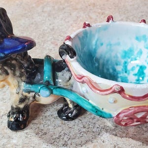 Może przedstawiać: Ceramiczna figurka osła ciągnąca ozdobny wózek. Osioł ma niebieski kapelusz i turkusową uprząż. Wózek jest biały z czerwonymi i turkusowymi akcentami.