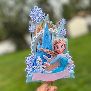 May include: A multi-layered cake topper featuring a princess in a blue dress, a castle, and a snowman. The topper has blue and white snowflake accents and a light pink banner. The overall design is a 3D effect.