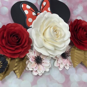 May include: A paper flower arrangement featuring a large white rose, two red roses, and several smaller pink and black flowers. The arrangement is accented with gold leaves and a black and red Minnie Mouse bow.