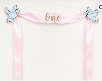 May include: A pink satin ribbon banner featuring the word One in gold glitter script. The banner is flanked by two light blue cartoon bird cutouts, each attached to the ends of the ribbon, with two long pink satin ribbons hanging vertically downward.