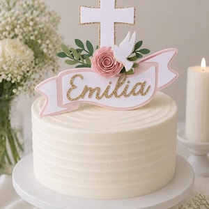 May include: A white frosted cake on a white cake stand, topped with a decorative cross, banner, and floral arrangement. The banner reads "Emilia" in gold glitter lettering. A white dove and pink rose are also featured.