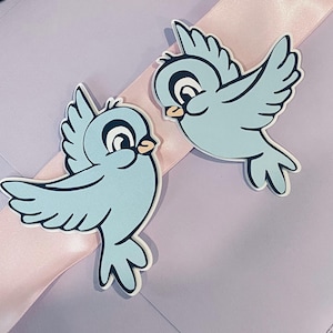May include: Two light blue bird-shaped cutouts with black and white details. The birds are positioned on a light purple surface with a pink ribbon. The birds have a cartoonish design with open wings.