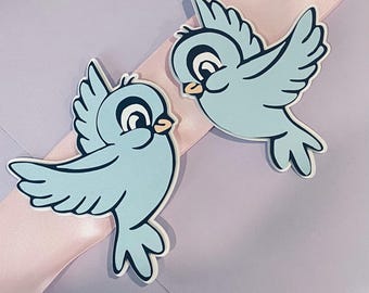 May include: Two light blue bird-shaped cutouts with black and white details. The birds are positioned on a light purple surface with a pink ribbon. The birds have a cartoonish design with open wings.