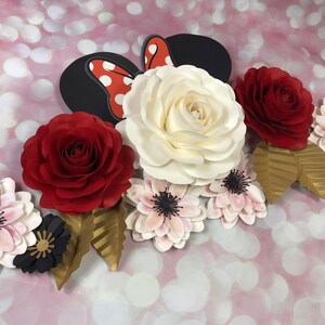 May include: A paper flower arrangement featuring a large white rose, two red roses, and several smaller pink and black flowers. The arrangement also includes gold leaves and a red and white polka dot bow.