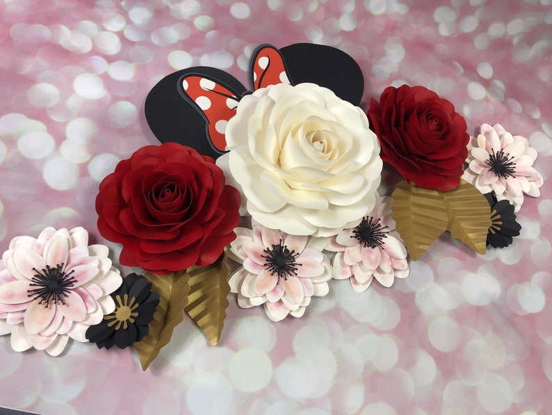 May include: A paper flower arrangement featuring a large white rose, two red roses, and several smaller pink and black flowers. The arrangement also includes gold leaves and a red and white polka dot bow.
