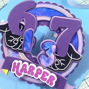 May include: A purple and blue birthday cake topper with the name "Harper" and the numbers "673". The topper has a layered design with a banner and decorative elements. The background is light blue.