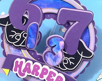 May include: A purple and blue birthday cake topper with the name "Harper" and the numbers "673". The topper has a layered design with a banner and decorative elements. The background is light blue.