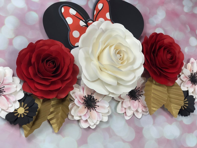 May include: A paper flower arrangement featuring a large white rose, two red roses, and several smaller pink and black flowers. The arrangement is accented with gold leaves and a black and red Minnie Mouse bow.