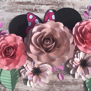 May include: A set of paper flowers in shades of pink and white, with a black and red Minnie Mouse ears cutout in the center. The flowers are arranged in a semi-circle, with the Minnie Mouse ears in the center.