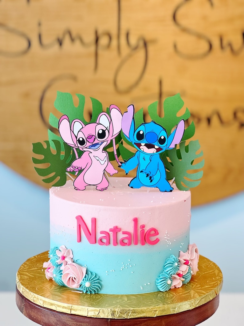 May include: A two-tiered cake decorated with blue and pink fondant figures of Stitch and Angel from the Disney movie Lilo & Stitch. The cake is decorated with pink and blue frosting and has the name "Natalie" written in pink frosting on the front.